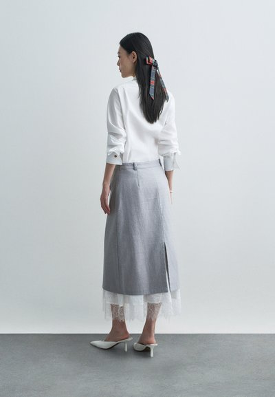 Balta marškiniai su sukneštais rankovėmis ir mygtuko detale gale, derinama su pilka sijonu, turinčiu nėrinių apdailą ir šoninius plyšius, dėvima su baltomis aukštakulnėmis bateliais.