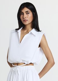 Blusa bianca senza maniche con scollatura a V, dotata di una vestibilità ampia e orlo arricciato, realizzata in tessuto morbido; texture liscia e dettagli minimi.