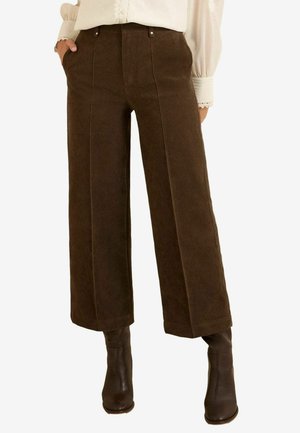 Kobieta ubrana w ciemnobrązowe spodnie typu szerokie nogawki, kremową bluzkę z koronkowymi mankietami oraz ciemnobrązowe botki za kostkę, z rękami w kieszeniach.