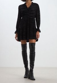 Svart randig långärmad klänning med knäppt halsringning, utsvängd nederdel, i kombination med höga svarta stövlar som går upp till låren och med en styv klack.