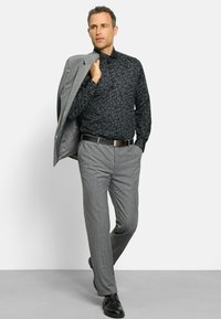 Schwarzes gemustertes Hemd mit langen Ärmeln, graue Anzugjacke über einer Schulter, graue Hose und schwarze Dressschuhe.