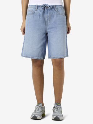 Person iført lyseblå denimshorts med bindebånd i taljen, hvid top og grå atletiksko, stående mod en ensfarvet baggrund.