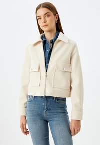 Gekürzte cremefarbene Jacke mit zwei vorderen Taschen, einem Reißverschluss und einem Kragen, getragen über einem blauen Jeanshemd und kombiniert mit blauen Jeans.