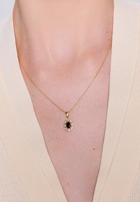 Złoty naszyjnik z owalnym czarnym kamieniem otoczonym małymi przezroczystymi kamieniami, spoczywający na skórze nad jasną, teksturowaną odzieżą.