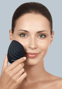 Dispositif de nettoyage du visage noir avec un design incurvé et en couches tenu contre la joue d'une femme. Texture en plastique lisse, forme ergonomique et logo visible.