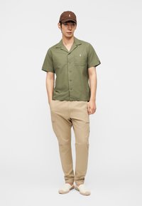 Mees, kes kannab pruuni nokamütsi, oliivirohelisest lühikeste varrukatega nööbitavat särki rinnataskuga, beežikaid taskupükse ja valgeid libisevaid kingi, seisab.