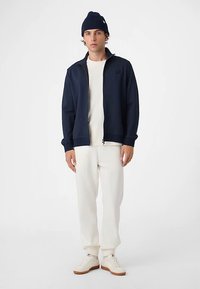 Marineblå ribstrikket lynlåstrøje med høj krave, båret over en hvid sweater og cremefarvede bukser. Suppleret med en marineblå beanie og hvide sneakers.