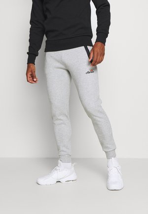 Persoon draagt lichtgrijze joggingbroek met zwarte ritszakken en witte sneakers, staand tegen een egale lichte achtergrond.