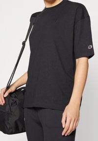 Černé bavlněné tričko s krátkými rukávy, žebrovaným kulatým výstřihem a malým logem vyšitým na hrudi a rukávu. Doplněno černou taškou.
