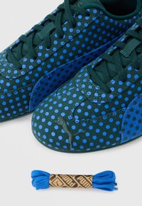 Baskets vertes Puma avec des pois bleus et des lacets, présentées avec une paire supplémentaire de lacets bleu vif en dessous.