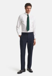 Witte overhemdbloes, donker groene en gestreepte stropdas, marineblauwe pantalon en donkerbruine leren schoenen; gladde stof en getailleerde pasvorm.