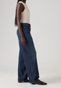 Kvinna som bär en ärmlös beige topp, höga vida mörkblå jeans och svarta lågklackade skor, stående på sidan mot en ljusgrå bakgrund.