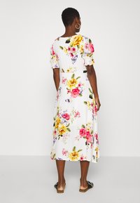 Blommig klänning i vitt, med sprudlande rosa, gula och lila blommor. Medellång med puffärmar och figurnära midja. Platta sandaler synliga.