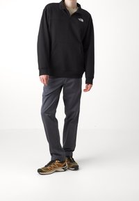 Pullover in pile nero con zip a quarto, tasca frontale e logo. Pantaloni grigi e scarpe da corsa con dettagli gialli completano l'outfit.
