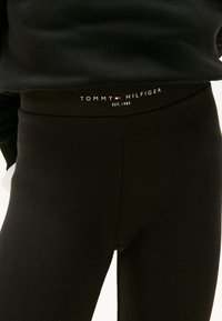 Pantalones negros ajustados con una etiqueta en la cintura que dice "Tommy Hilfiger EST. 1985" sobre una persona que lleva una camiseta negra.