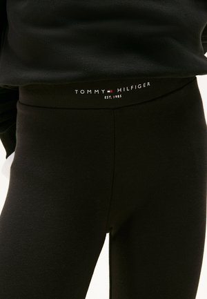 Pantalones negros ajustados con una etiqueta en la cintura que dice "Tommy Hilfiger EST. 1985" sobre una persona que lleva una camiseta negra.
