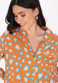 Orange skjorta med knappar och blåa samt rosa leopardfläckar, korta ärmar och en bröstficka. Tillverkad av lätt tyg med en slät textur.