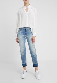 Blusa blanca de botones con cuello y detalles de pliegues, combinada con jeans desgastados de color azul claro que tienen puños más oscuros y zapatillas blancas.