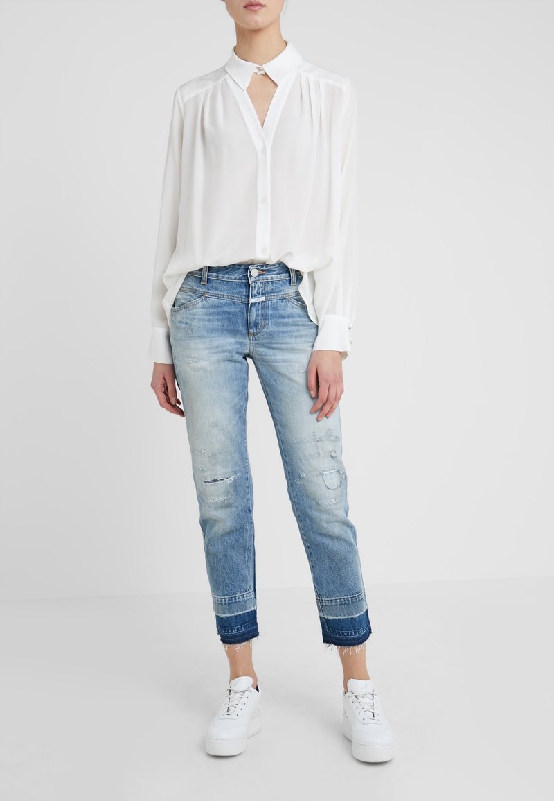 Blusa blanca de botones con cuello y detalles de pliegues, combinada con jeans desgastados de color azul claro que tienen puños más oscuros y zapatillas blancas.
