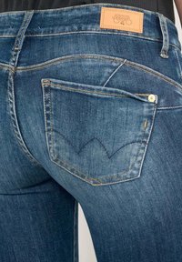 Jeans de mezclilla en un lavado azul medio, con un diseño de bolsillo trasero curvado, costuras doradas y un parche de piel de la marca en la cinturilla.