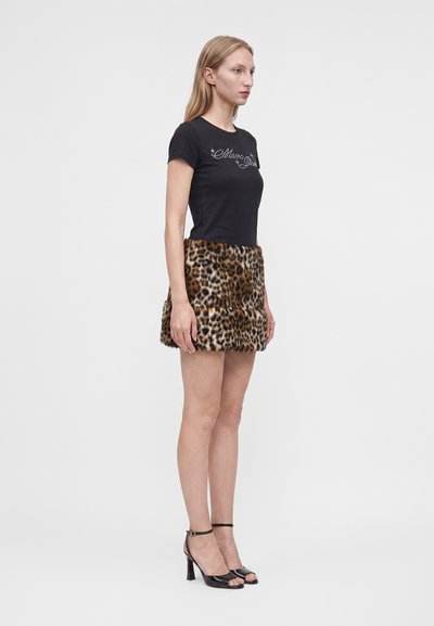 Femme debout de profil portant un t-shirt noir avec logo, une jupe courte à imprimé léopard et des sandales à talons noirs sur fond uni.