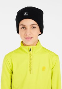 Beanie a coste nera con logo, abbinato a una maglia a maniche lunghe gialla brillante con mezza zip e branding stampato sulla parte anteriore.