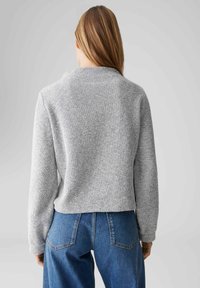 Donna con capelli lunghi castano chiaro che indossa un maglione grigio chiaro texturizzato e jeans blu a vita alta, vista da dietro su uno sfondo semplice.