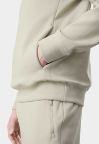 Jasno beżowy sweatshirt z boczną kieszenią i �ściągaczami. Materiał wydaje się miękki i gładki, z detalem logo po boku.