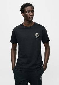 Μαύρο βαμβακερό μπλουζάκι με κοντά μανίκια, με λευκό κεντητό σχέδιο κάκτου σε γλάστρα στο αριστερό στήθος.