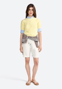 Lichtgele gebreide trui met korte mouwen, witte shorts en een panterprint sjaal om de taille geknoopt. Neutrale flip-flops maken de look compleet.