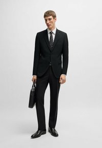 Completo nero con giacca aderente e pantaloni a gamba dritta. Camicia bianca e cravatta a righe. Scarpe in pelle nere e una valigetta coordinata.