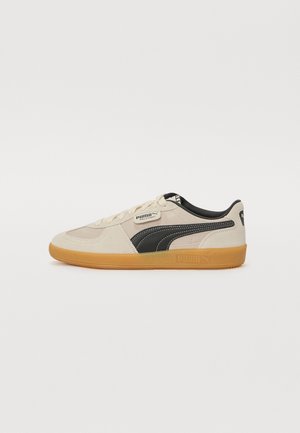 Beige en zwarte Puma Palermo sneaker met gumzolen, zijlogo-streep, vetersluiting en gevoerde enkelkraag op een witte achtergrond.