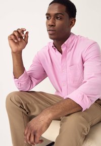 Vaaleanpunainen nappipaita rintataskulla, pitkät hihat käännettynä kalvosimista. Yhdistetty vaaleisiin khakinvärisiin housuihin, kangas näyttää sileältä.