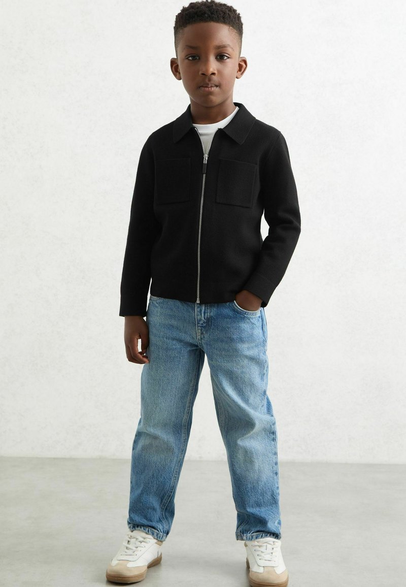 Veste zippée noire avec deux poches avant sur une chemise blanche, associée à un jean en denim bleu clair et des baskets blanches avec des accents beiges.