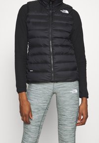 Schwarze Steppweste mit Reißverschluss und hohem Kragen. Getragen über einem schwarzen Langarmoberteil, kombiniert mit strukturierten grauen Leggings mit einem Logo.