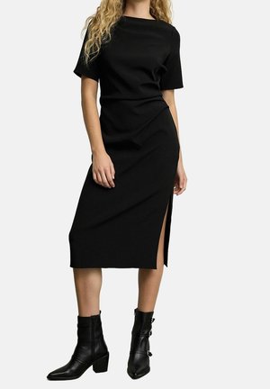 Femme portant une robe midi noire à manches courtes avec une fente latérale, associée à des bottines noires à talons moyens avec boucles.