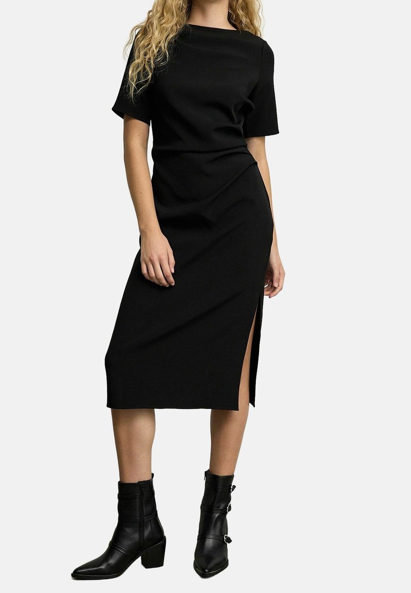 Femme portant une robe midi noire à manches courtes avec une fente latérale, associée à des bottines noires à talons moyens avec boucles.
