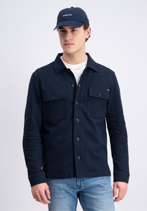 Junger Mann trägt ein marineblaues Hemd mit Knöpfen und zwei Brusttaschen, hellblaue Jeans, weißes Unterhemd und eine marineblaue Kappe mit weißem Schriftzug.