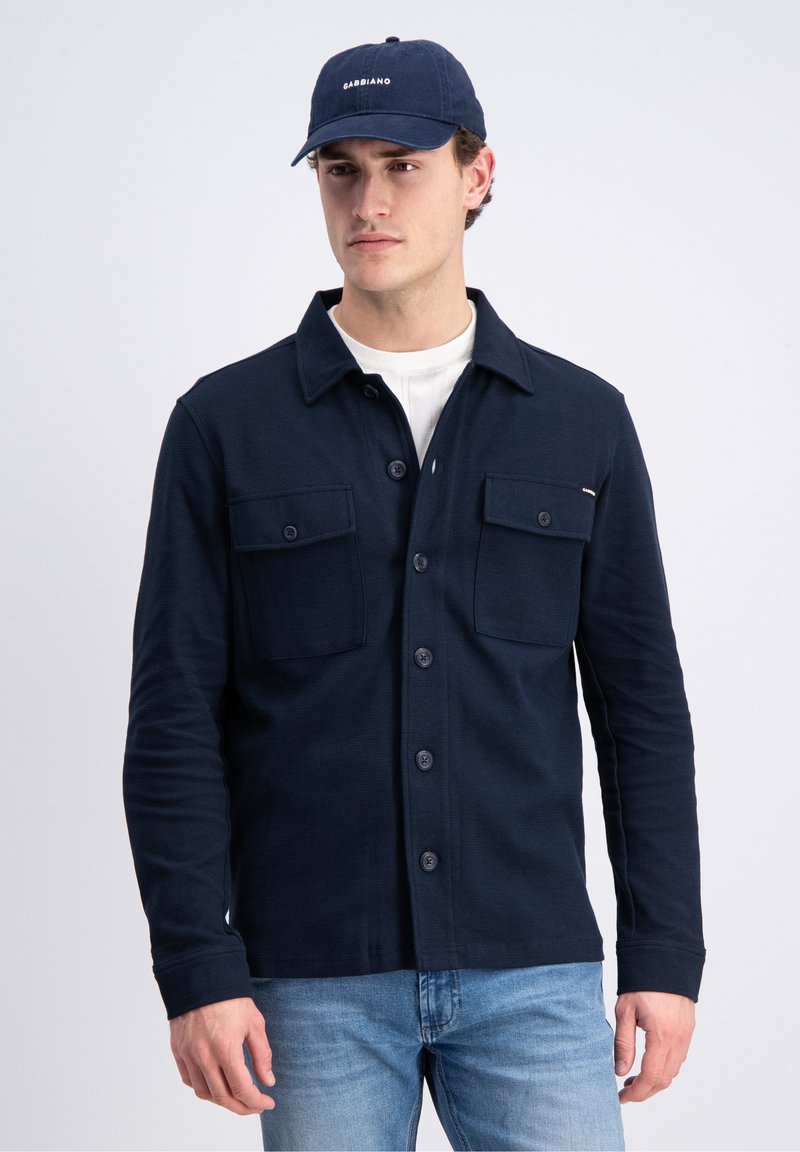 Jonge man met een marineblauw overhemd met knopen en twee borstzakken, lichtblauwe spijkerbroek, witte ondershirt en een marineblauwe pet met witte tekst.