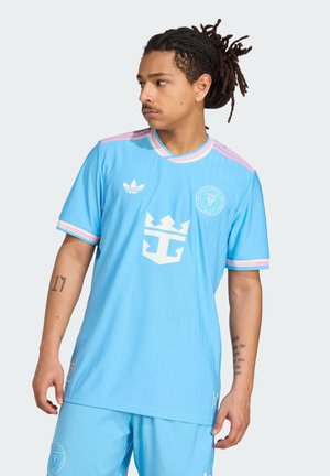 Maglia da calcio azzurro chiaro con accenti rosa, caratterizzata da un colletto tondo, strisce verticali e un grande logo bianco sul petto.
