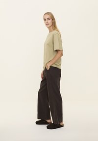 Világos olajzöld pamut póló, laza fazonban, sötét, széles szárú nadrággal és fekete slip-on cipővel párosítva, minimalista dizájn.