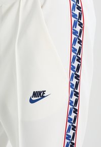 Vitaid sportbyxor med blå Nike-logotyp, med en sidremsa i blå, röd och svart mönster. Slät textur, smal passform.