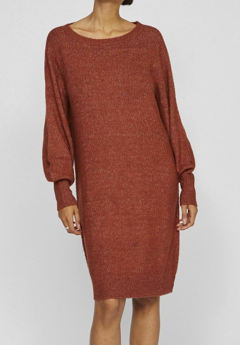 Robe pull en maille rouge rouille, dotée d'une coupe ample, d'un large col bateau et de manches longues bouffantes avec des poignets côtelés. Texture douce.