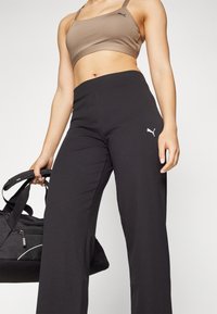 Čierne rebrované nohavice s širokými nohavicami a elastickým pásom, na boku s logom Puma v bielej farbe; model drží čiernu športovú tašku.