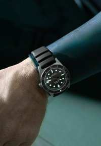 Muñeca con un reloj de buceo de goma negra que muestra manecillas y marcadores luminosos, llevado sobre una manga de traje de neopreno color teal.