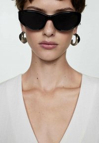 Schwarze Katzenaugen-Sonnenbrille, silberne Creolen und ein weißes V-Ausschnitt-Oberteil. Glatte Hauttextur mit dezenten Sommersprossen sichtbar.