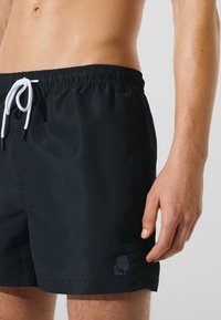 Črne kopalne kratke hlače iz lahkega materiala, z elastičnim pasom z vrvico za zategovanje in majhnim logotipom na spodnjem levem delu.