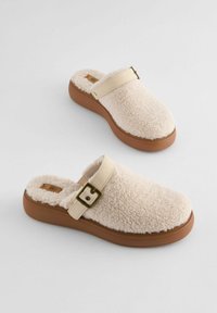Sabots blancs pelucheux avec une semelle en caoutchouc beige et une sangle beige comportant un détail de boucle en métal. Texture douce sur l'ensemble.