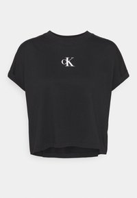 Camiseta negra corta de manga corta con puños enrollados y logo blanco "cK" centrado en el pecho, sobre un fondo liso.
