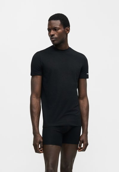 Άνδρας που φορά στενό μαύρο κοντομάνικο πουκάμισο και μαύρα μπόξερ, στέκεται σε απλό ανοιχτό φόντο, κοιτάζοντας στο πλάι.